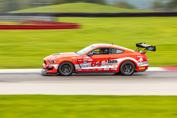 Ein Ford Mustang GT Rennauto, verziert mit Text und Zahlen, fährt auf einer Rennstrecke mit einem Helmträger im Sitz, der Hintergrund ist unscharf, um Bewegung zu vermitteln.