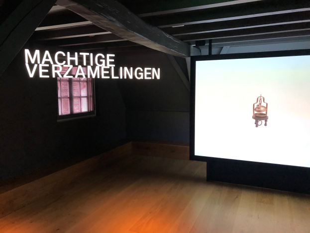 Installationsansicht der Ausstellung 'Machtige Sammlungen' im Museum of Modern Art in Berlin, mit einem großen Bildschirm auf der rechten Seite und Text an der Wand dahinter sowie einem Fenster auf der linken Seite.