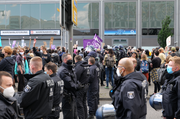 Große Gruppe von Menschen demonstriert vor einem Gebäude, einige halten Schilder und tragen Helme, mit einem Schildständer und einem Baum im Vorder- und Hintergrund.