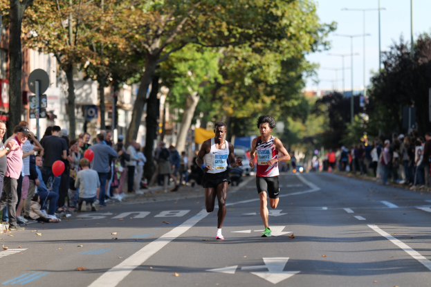 Zwei Männer bei einem Marathon auf einer Stadtstraße mit Zuschauern auf der linken Seite und Gebäuden, Bäumen und einem klaren Himmel im Hintergrund.