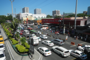 Eine belebte Stadtstraße mit verschiedenen Fahrzeugen, Fußgängern, Gebäuden und einem klaren blauen Himmel.