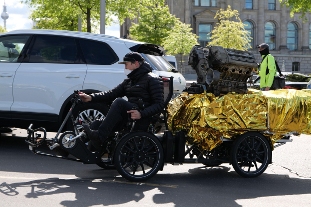 Ein Mann in einem Rollstuhl mit einem großen Motor auf dem Rücken, umgeben von Fahrzeugen auf einer Straße mit Bäumen, Gebäuden, Polen und einem klaren blauen Himmel im Hintergrund; er trägt eine schwarze Jacke, eine Mütze und hält ein Objekt in der Hand.