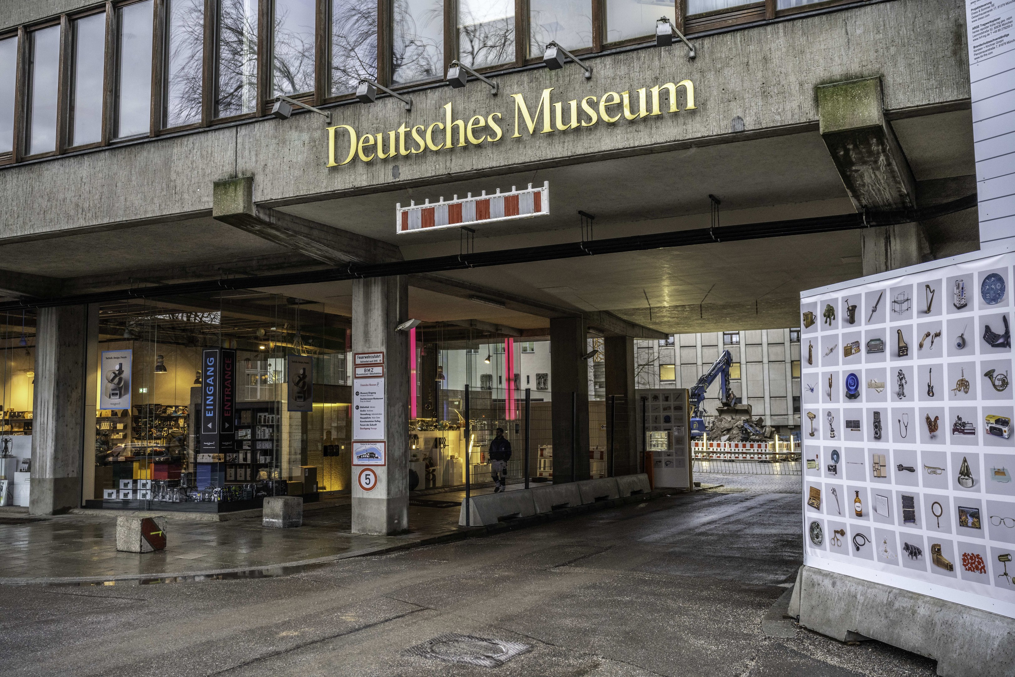 Außensicht des Deutschen Museums in Berlin: Ein großer Bau mit Glasfenstern, Säulen und einem Namensschild, neben einer Informationsanzeigetafel, mit Bäumen, anderen Gebäuden und Himmel im Hintergrund.