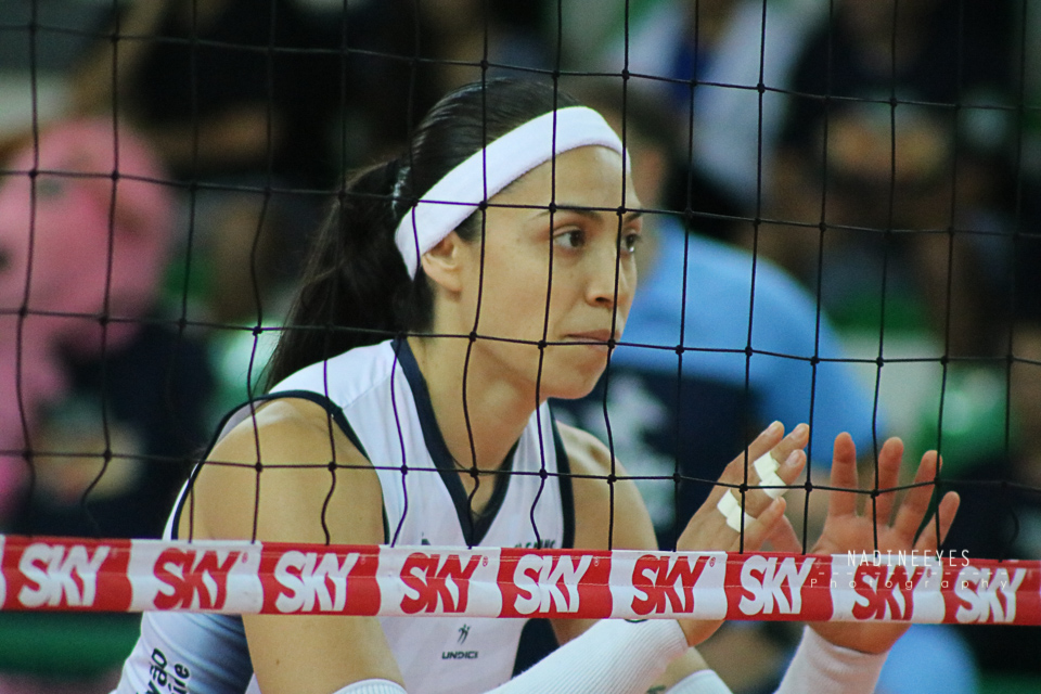 Eine weibliche Volleyballspielerin in Uniform mit entschlossener Miene ist auf dem Feld mit einem Netz im Vordergrund und einem unscharfen Hintergrund im Einsatz.