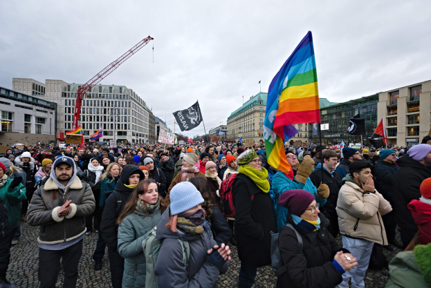 Große Gruppe von Menschen bei einer LGBTQ+-Rechte-Demonstration in Berlin, die Fahnen und Transparente schwenken, mit Gebäuden, einem Kran und einem bewölkten Himmel im Hintergrund.