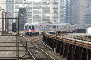 Nahverkehrszug fährt neben Hochhäusern auf erhöhten Gleisen mit sichtbarer Bahninfrastruktur und städtischen Elementen im Hintergrund.