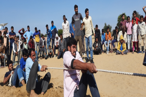 Eine Gruppe von Menschen, die auf einem sandigen Strand sitzen, mit einem Mann, der ein Seil in der Vordergrund hält, Bäume, Gebäude und einen klaren blauen Himmel im Hintergrund und ein Tuch, das über einen Stuhl auf der rechten Seite drapiert ist, was auf einen Seilziehen-Wettbewerb hindeutet.