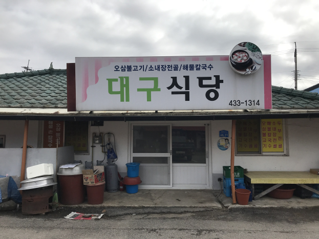 Kleines Gebäude mit einer Tafel, die 'Koreanisches Restaurant' lautet, eine Tür, Fenster, Behälter, Eimer, ein Tisch, Pfosten, Drähte, ein weiteres Haus und ein bewölkter Himmel.