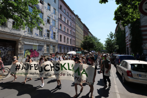 Eine Gruppe von Menschen marschiert auf einer Straße in Berlin, hält ein Transparent, mit parkenden Autos auf der rechten Seite, einem Radfahrer auf der linken Seite und Gebäuden, Bäumen und einem Schild im Hintergrund unter einem sichtbaren Himmel.