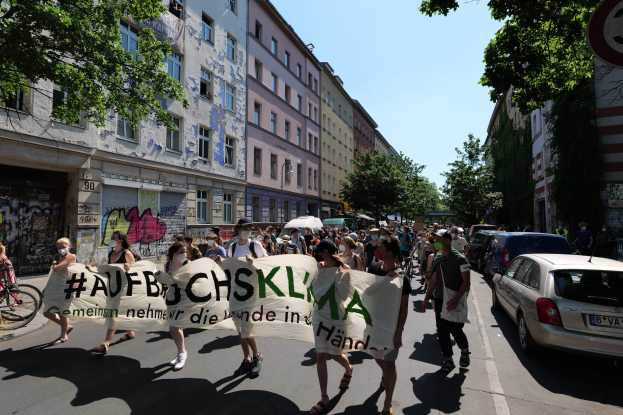 Eine Gruppe von Menschen marschiert auf einer Straße in Berlin, hält ein Transparent, mit parkenden Autos auf der rechten Seite, einem Radfahrer auf der linken Seite und Gebäuden, Bäumen und einem Schild im Hintergrund unter einem sichtbaren Himmel.