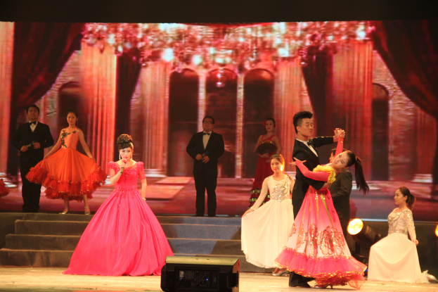 Eine Gruppe von Menschen auf einer Bühne mit Stufen und einem Bildschirm im Hintergrund, beleuchtet von Scheinwerfern, mit einem Lautsprecher unten, was eine Tanzvorstellung suggeriert.