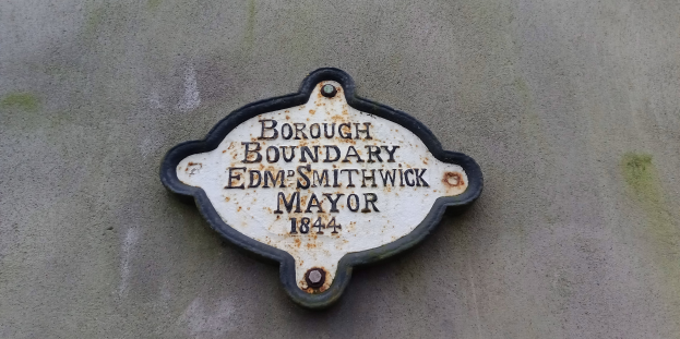 Metallschild an einer Gebäudeseite, das die Borough-Grenze markiert, mit "Borough-Grenze Edm Smithwick Mayor" in fetten weißen Buchstaben auf einem schwarzen Hintergrund.