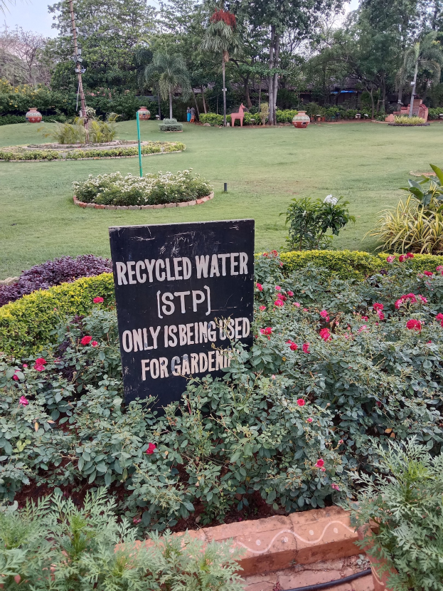 Ein Garten mit einem Schild, auf dem "Recycled Water (STP) Only Being Used for Gardening" steht, umgeben von grünem Gras, bunten Blumen, hohen Bäumen und einem klaren blauen Himmel.