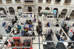 Eine große Gruppe von Menschen auf einer internationalen Fachmesse mit Ständen, Bannern, Tischen, Stühlen, Luftballons und einem Gebäude mit Bögen, Säulen, Geländern und einem Bildschirm im Hintergrund.