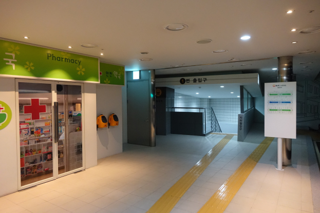 Apotheken-Eingang mit einem "Apotheke"-Schild auf der linken Seite, einer Tafel mit Text auf der rechten Seite, Hintergrundobjekten und beleuchtetem Dach.