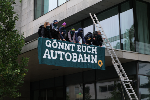 Eine Gruppe von Menschen auf dem Dach eines Gebäudes, die eine Fahne halten, mit einem Baum links und Glasfenstern im Hintergrund, wahrscheinlich protestieren sie für die deutsche Regierung.