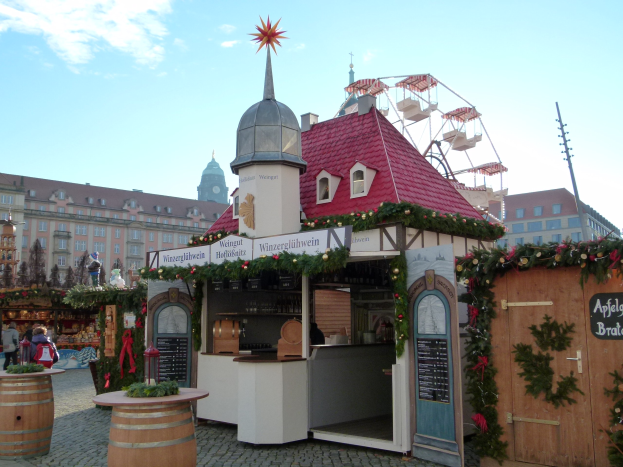 Kleines rotes Dachgebäude mit einem Riesenrad davor, umgeben von Festdekorationen, Menschen und Gegenständen, vor einem Hintergrund aus Gebäuden, Bäumen und einem bewölkten Himmel auf einem Oktoberfest in München, Deutschland.