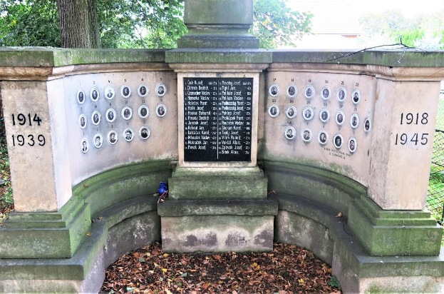 Denkmal im jüdischen Friedhof in Berlin, Deutschland, mit einer Text- und Nummerntafel an seiner Wand, umgeben von Bäumen, einem Zaun und trockenen Blättern auf dem Boden.
