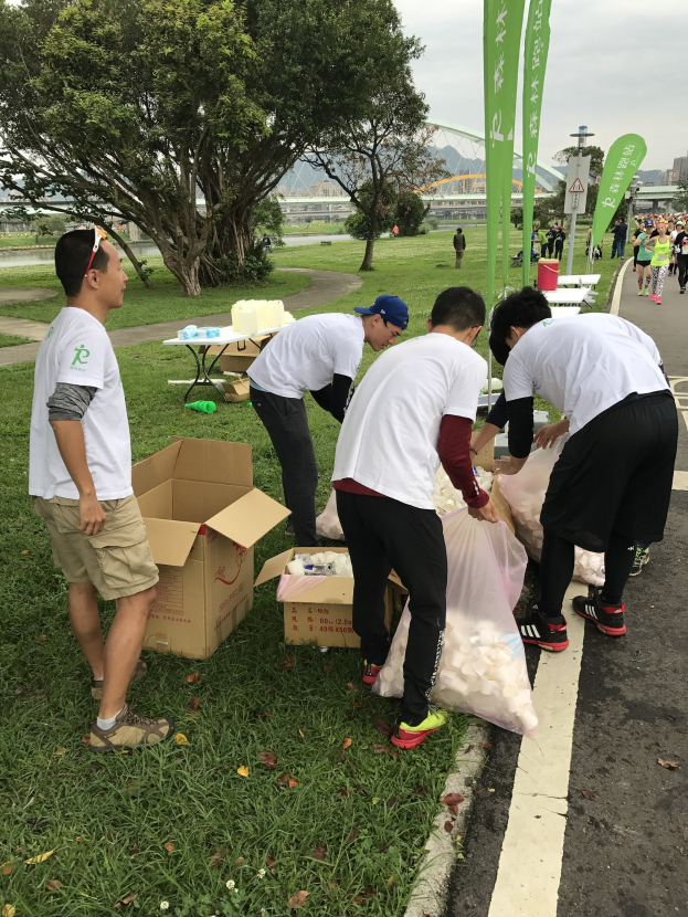 Group of people on a roadside with scattered cardboard boxes and bottles, participating in a charity run with banners visible in the background.