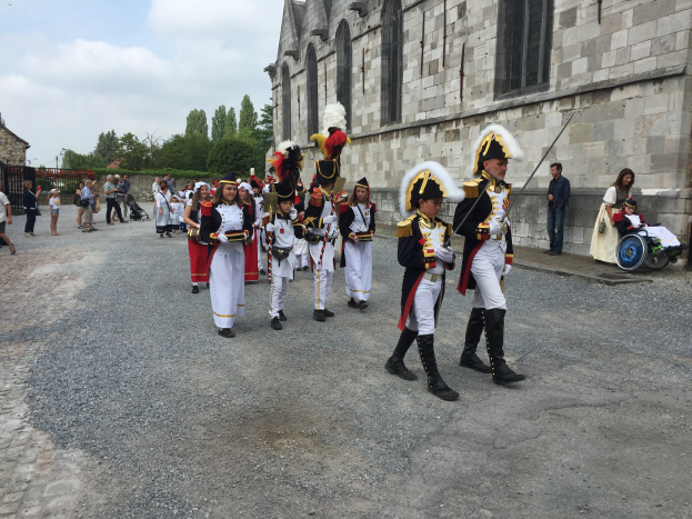 Eine Gruppe uniformierter Personen, einige im Rollstuhl und andere mit Gewehren, marschiert auf einer Straße in einem Militärumzug in Frankreich, mit einem Gebäude, Bäumen und einem bewölkten Himmel im Hintergrund.