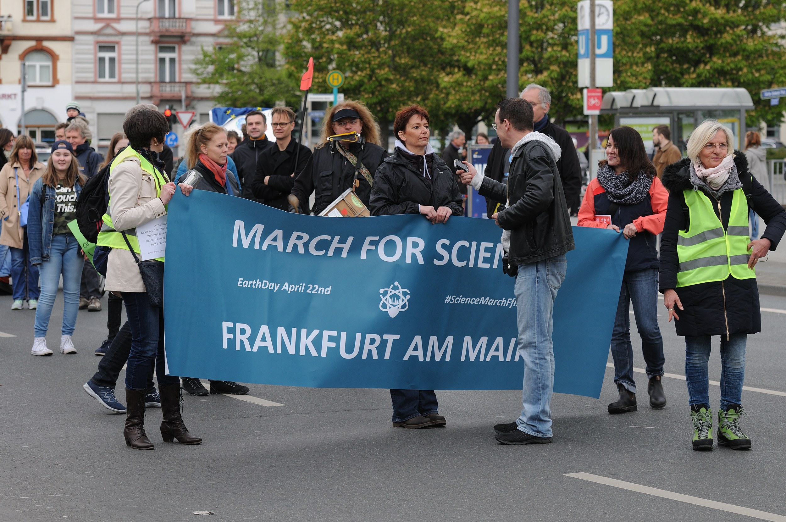 Eine bunte Gruppe von Menschen marschiert auf einer Straße, die eine 'March for Science Frankfurt am Main'-Fahne hält, mit Bäumen, Gebäuden und einem klaren Himmel im Hintergrund.