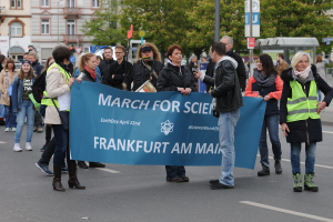 Eine bunte Gruppe von Menschen marschiert auf einer Straße, die eine 'March for Science Frankfurt am Main'-Fahne hält, mit Bäumen, Gebäuden und einem klaren Himmel im Hintergrund.