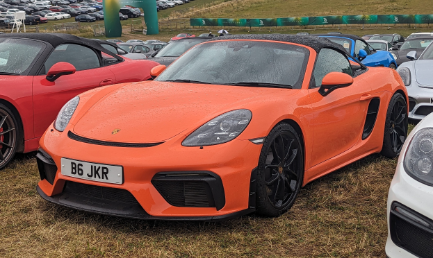 Roter Porsche 718 Boxster GTS auf dem Goodwood Festival of Speed mit Autos, Bannern, Bäumen und einem klaren blauen Himmel im Hintergrund.