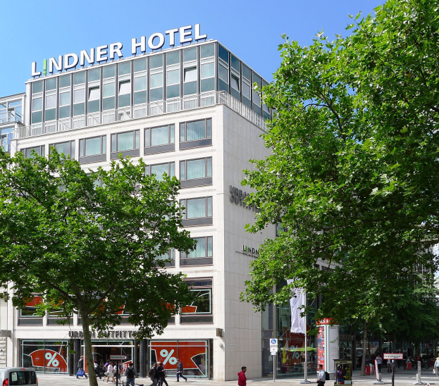 Großes modernes Gebäude mit der Aufschrift "Lindner Hotel" in Berlin, mit Glasfenstern, umgeben von Bäumen und einer belebten Straße mit Fussgängern und Fahrzeugen.