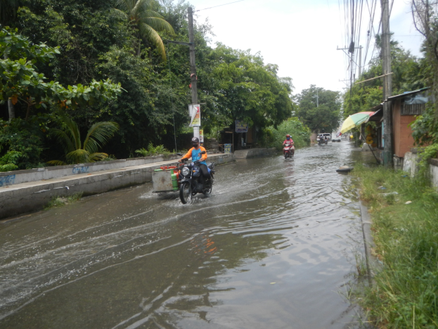 Eine Gruppe von Menschen fährt mit Motorrädern durch eine überflutete Straße, umgeben von Vegetation, Gebäuden und Strommasten, mit einem klaren Himmel im Hintergrund.