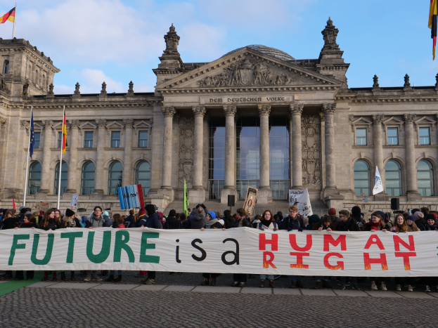 Eine Gruppe von Menschen hält ein Transparent mit der Aufschrift 'Zukunft ist ein Menschenrecht' vor dem Reichstagsgebäude in Berlin, Deutschland, mit sichtbaren Fenstern, Säulen und Bögen des Gebäudes, umgeben von Fahnenmasten, unter einem bewölkten Himmel.