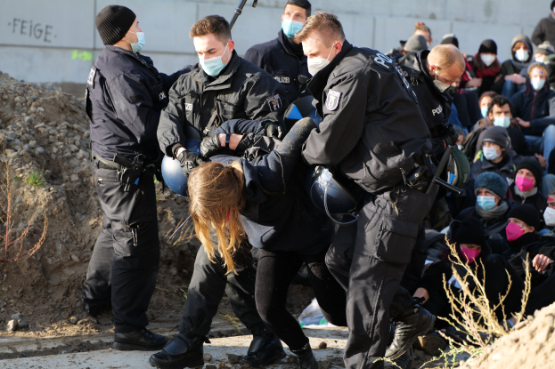 Eine Gruppe maskierter Personen in Kapuzenpullis, einige bewaffnet, umringt eine Frau, die von der Polizei eskortiert wird, mit urbanen Elementen wie Pflanzen, Steinen und einer Wand mit Protesttext im Hintergrund.