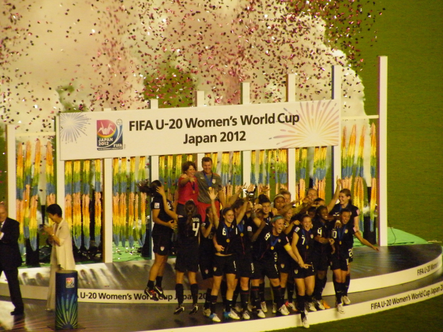Gruppe von Frauenfußballspielerinnen in schwarzer Uniform, die auf einer Bühne feiern, umgeben von einer Menge mit Kameras, mit einem dekorierten Hintergrund und grasbewachsenem Boden.