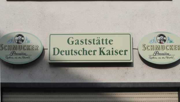 Drei hölzerne Schilder mit der Aufschrift "Gaststätte Deutscher Kaiser Schmucker" an einer Gebäudewand über einer geschlossenen Jalousie angebracht.