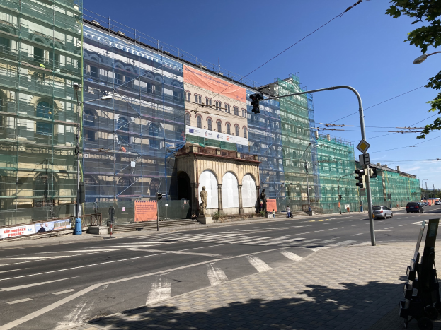Großer mehrstöckiger Bau unter construction in Berlin, umgeben von Straßeninfrastruktur, Fahrzeugen, Fußgängern und Bäumen unter einem bewölkten Himmel.