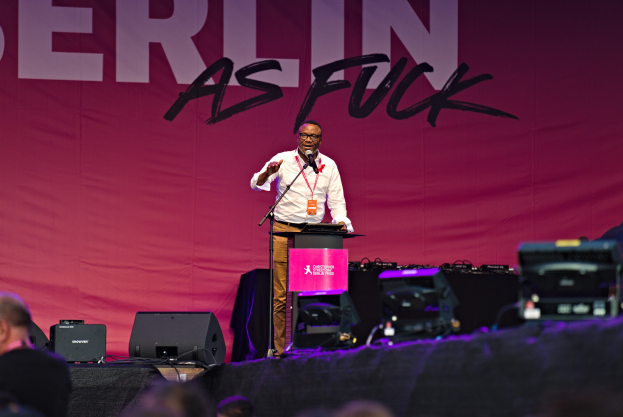 Ein Mann in einem weißen Hemd und braunen Hosen steht auf einer Bühne und hält ein Mikrofon in der Hand, vor einem pinken Banner mit der Aufschrift "Berlin as Fuck" und sichtbaren Lautsprechern auf der Bühne.