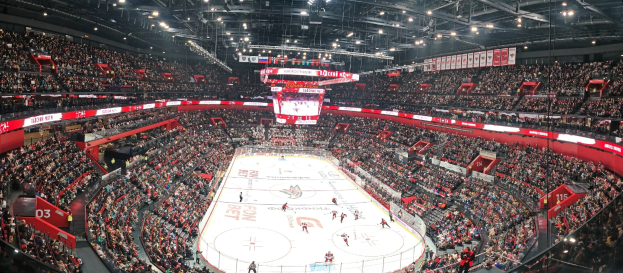 Eine große Indoor-Hockey-Arena mit zahlreichen Zuschauern unter hellen Deckenlampen, einem zentralen Scoreboard und einer enthusiastischen Menge.