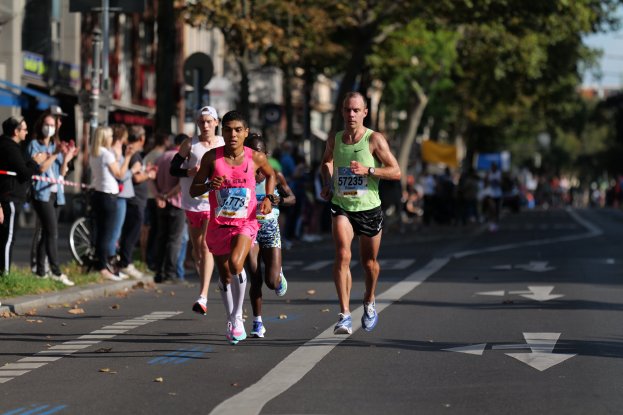 Gruppe von Menschen, die bei einem Marathon auf einer Stadtstraße laufen, mit Zuschauern auf der linken Straßenseite, Gras am Boden, Bäumen, Gebäuden, Polen, Brettern und einem Fahrrad im Hintergrund.