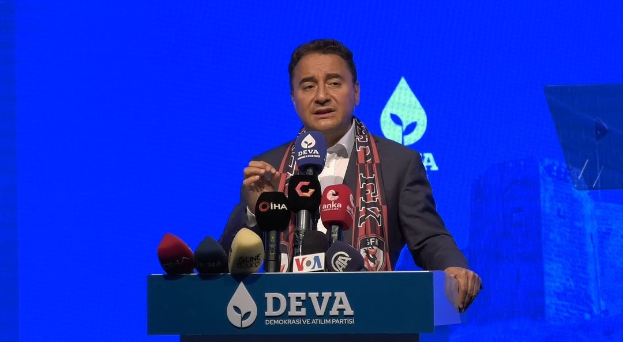 Mann in Mantel und Schal steht hinter Podium mit Mikrofonen und spricht hinter einer Tafel mit Text, der "deva ceo sagt, dass es keine gute Idee für die Zukunft ist", mit einem Bildschirm und einem Objekt im Hintergrund.