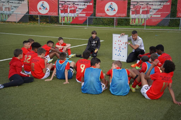 Junge Jungs auf einem Fußballfeld mit einer Tafel vor ihnen, einer den Fußball haltend, mit Bannern und Pflanzen im Hintergrund.