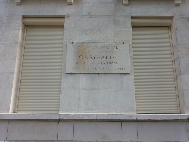 Ein zweistöckiges Gebäude mit zwei Fenstern, jeweils mit Läden, und einer Tafel an der Seite, wahrscheinlich ein Denkmal.