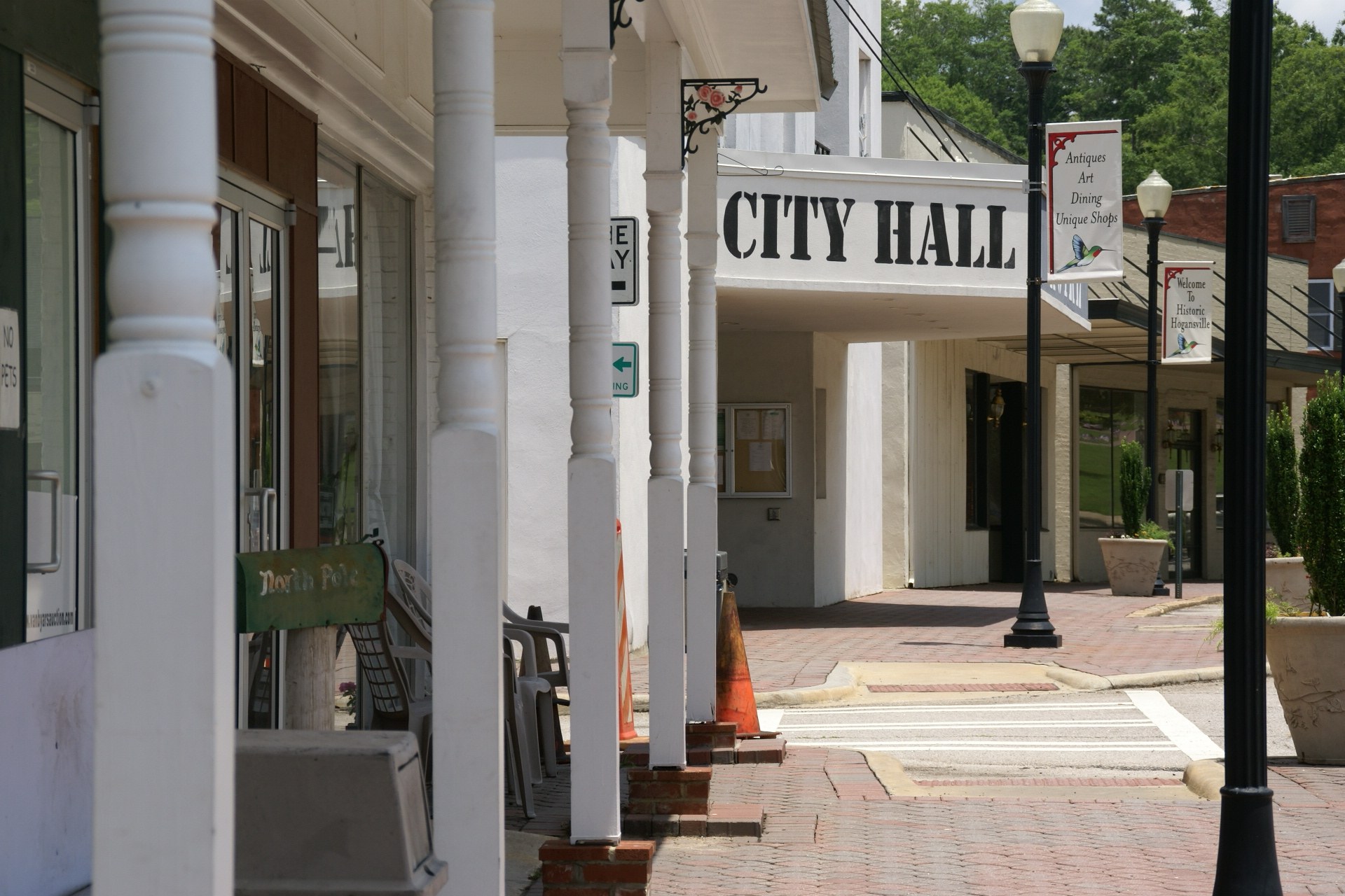 Stadthaus mit Gebäuden, Straßenschildern, Straßenlaternen, Namensschildern, Kraftfahrzeugen, Absperrgittern, Stühlen, Topfpflanzen, Bäumen und einem bewölkten Himmel im Hintergrund.