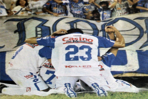 Gruppe von Fußballspielern auf dem Boden beim Feiern eines Tores vor einer jubelnden Menge, mit einem Banner mit Text im Hintergrund.