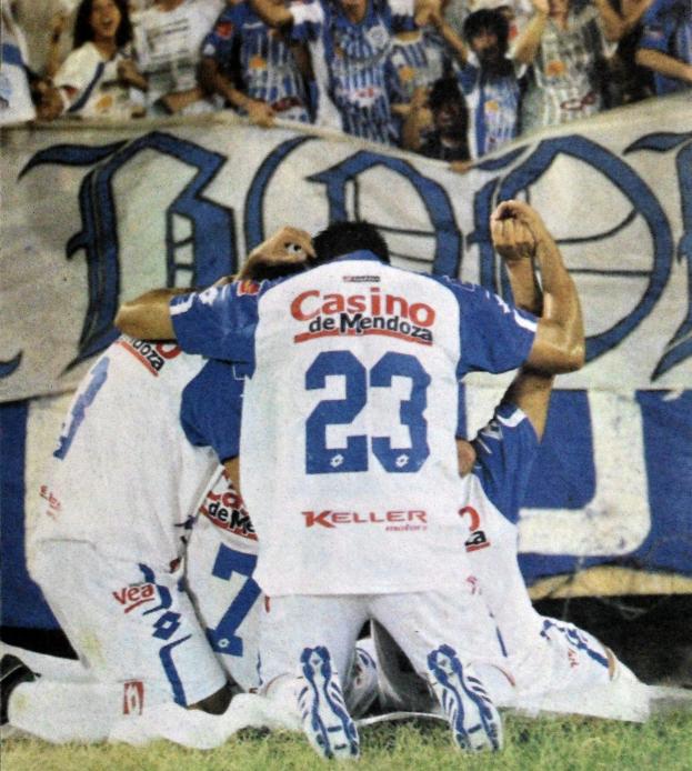 Gruppe von Fußballspielern auf dem Boden beim Feiern eines Tores vor einer jubelnden Menge, mit einem Banner mit Text im Hintergrund.