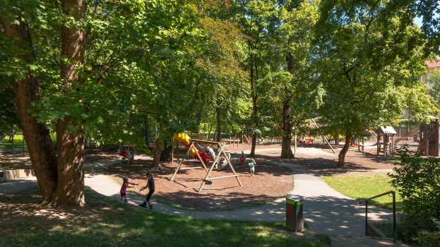 Ein Park mit einem Spielplatz in der Mitte, umgeben von Bäumen, mit Kindern auf der Ausrüstung, einem Mülleimer, Geländern, Pflanzen, Gras und Gebäuden im Hintergrund unter einem klaren blauen Himmel.