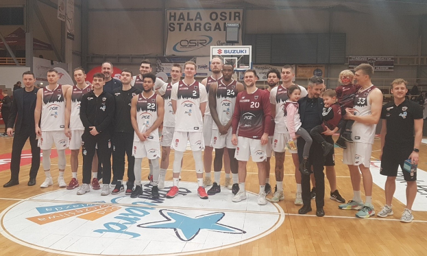 Mannergruppe auf einem Basketballfeld mit Kindern, Zuschauern, Bannern mit "Hala Osir Starga", einem Basketballkorb, Metallstangen, Deckenlampen und einer Wand im Hintergrund.