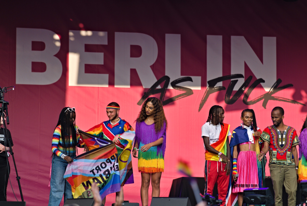 Eine Gruppe von Menschen auf einer Bühne mit einer Frau, die eine Flagge in der Mitte hält, einer Person, die eine Kamera auf einem Stativ links bedient, und einem Banner im Hintergrund mit der Aufschrift 'Berlin als Fuck 2019'.