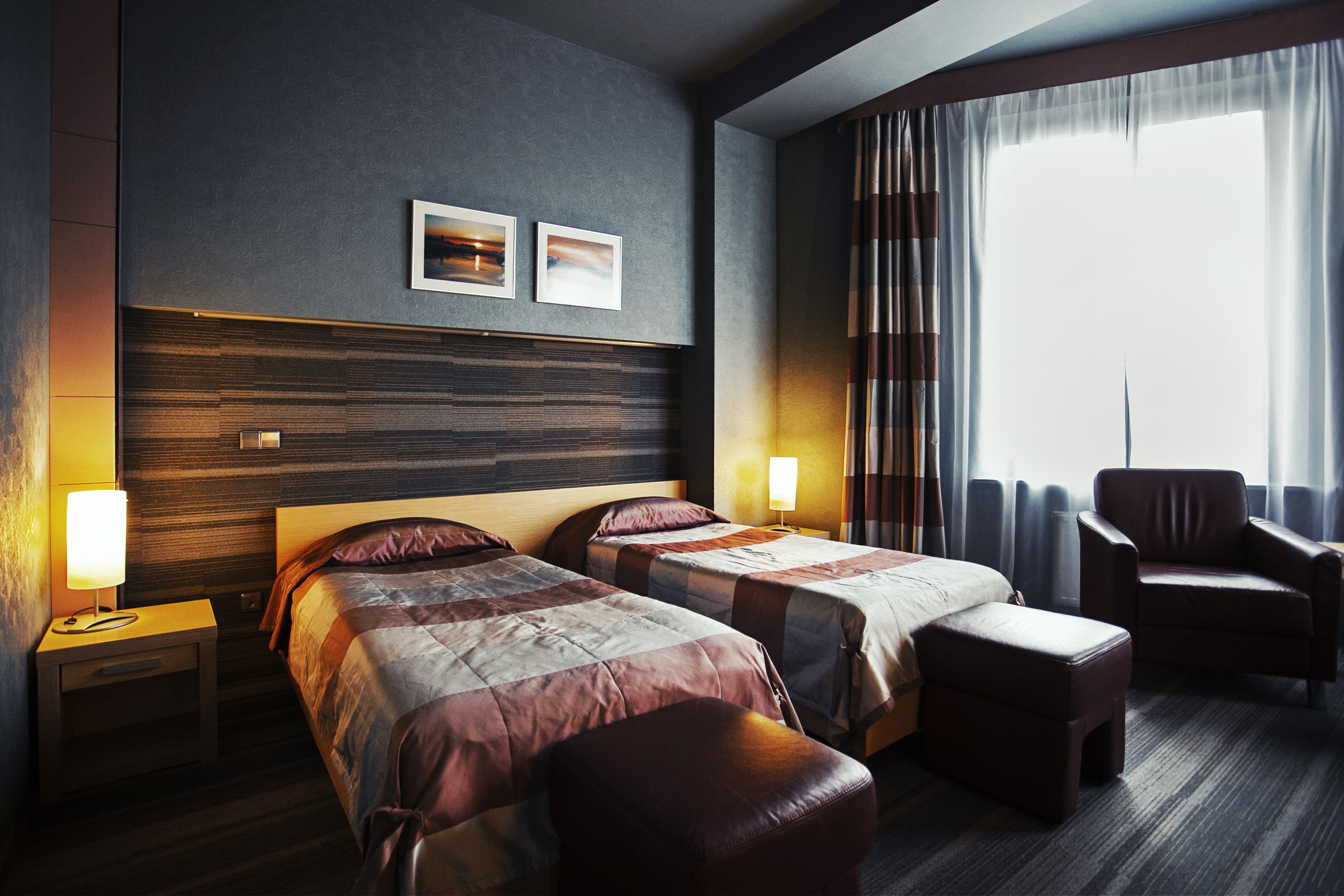 Hotelzimmer mit zwei Betten, jeweils mit Decken und Kissen, zwei Hockern, einem Stuhl, zwei Tischen mit Lampen, zwei gerahmten Fotos an der Wand und einem Fenster mit Vorhängen.