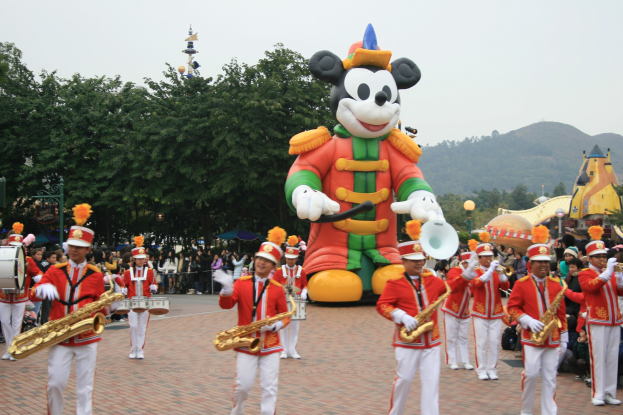 Eine Gruppe von Menschen in Kostümen spielt Musikinstrumente neben einem Mickey-Maus-Float in einem Umzug, mit Bäumen, Gebäuden, Hügeln und einem klaren blauen Himmel im Hintergrund; ein Schild mit Text ist links sichtbar.