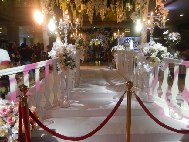 Eine Hochzeitszeremonie in einem Einkaufszentrum mit weißem Teppich, dekorativen Geländern, Blumenarrangements, Kronleuchtern und Deckenbeleuchtung, bei der eine Menge anwesend ist, darunter einige mit Taschen.