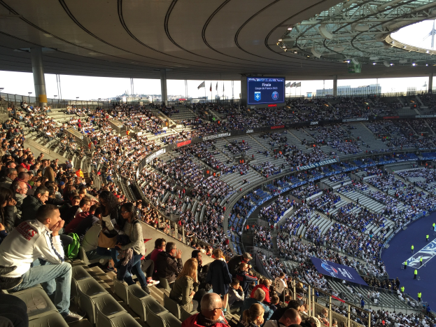 Große Menschenmenge in einem Stadion, die ein Fußballspiel auf einer Bühne mit Fahnen, Stangen und einem Bildschirm im Hintergrund schaut, identifiziert als Allianz Arena in München, Deutschland.